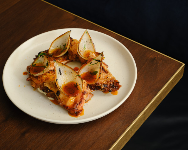 Grilled chicken wings are arranged on a white plate, topped with onion petals and drizzled with a reddish sauce, set on a wooden table with a gold-trimmed edge.