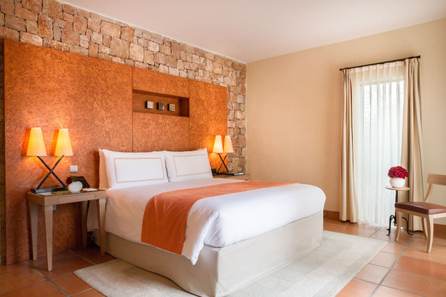 A neatly made bed with orange accents stands against a textured stone and orange wall. Two side tables hold lamps, surrounded by a beige room with a window draped in light curtains.