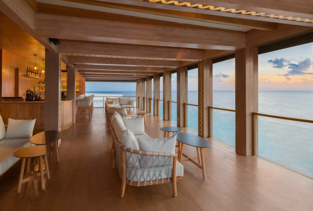 Bar lounge with chairs and tables lined up, offering a panoramic ocean view through large windows. The wooden interior creates a warm, inviting atmosphere enhanced by a sunset backdrop.