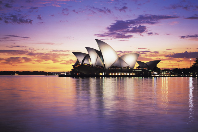 Iconic white sails of the Sydney Opera House are illuminated against a colorful sunset, reflecting on the calm harbor waters, creating a serene and picturesque scene