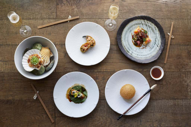 Five elegantly plated dishes rest on a wooden table, accompanied by chopsticks, glasses of champagne, and a small bowl of sauce, creating a sophisticated dining setting