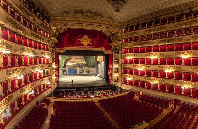 An opulent theater stage featuring a scenic backdrop is set for a performance, surrounded by ornate balconies with red velvet seats and golden decorations, viewed from the audience's perspective