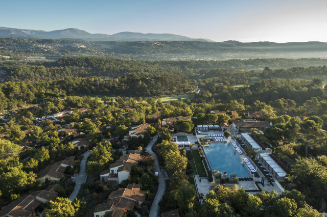 A large rectangular swimming pool sits surrounded by lounge chairs and umbrellas, amidst a cluster of buildings and trees, set against a backdrop of rolling hills and misty mountains.