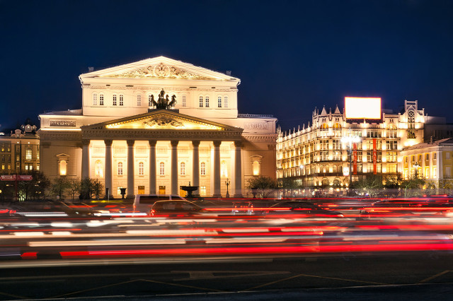 A grand, illuminated classical building stands at night, framed by a busy street with blurred car lights in the foreground, suggesting movement in an urban setting