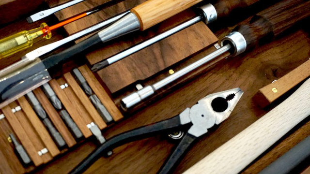 A set of woodworking tools, including chisels, a screwdriver, a set of drill bits, and pliers, are neatly arranged in a wooden toolbox