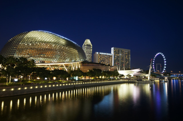 A modern waterfront cityscape at night features an illuminated domed building, Ferris wheel, and skyscrapers reflecting on the calm water, with trees lining the promenade
