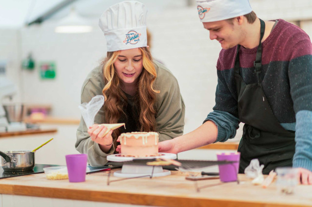 Two people decorate a cake together; one pipes icing while the other observes closely. They wear chef hats and aprons in a kitchen setting, surrounded by baking tools and ingredients.