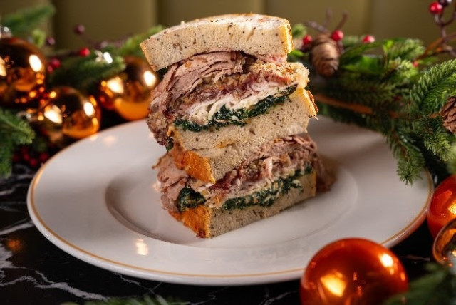 A stacked sandwich with turkey, greens, and stuffing sits on a white plate. It is surrounded by festive decorations, including golden ornaments and evergreen branches on a dark surface.