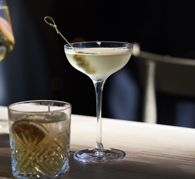 Martini glass with a yellow cocktail and olive on a light wood table.