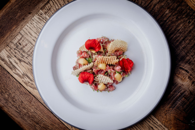 A gourmet dish with diced meat, waffle-cut potatoes, dollops of sauce, and red petals, artfully arranged on a white plate, placed on a wooden table.