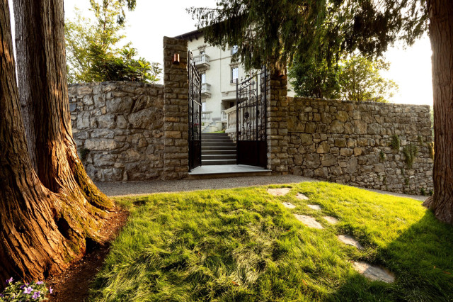A stone pathway leads to an open, black iron gate set into a tall stone wall. Lush grass and trees surround the entrance, with a house visible beyond.