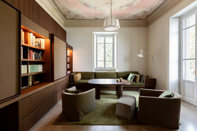 A cozy living room features brown armchairs and a central wooden table on a green rug, with bookshelves against the wall. A large window and decorative ceiling complete the elegant space.