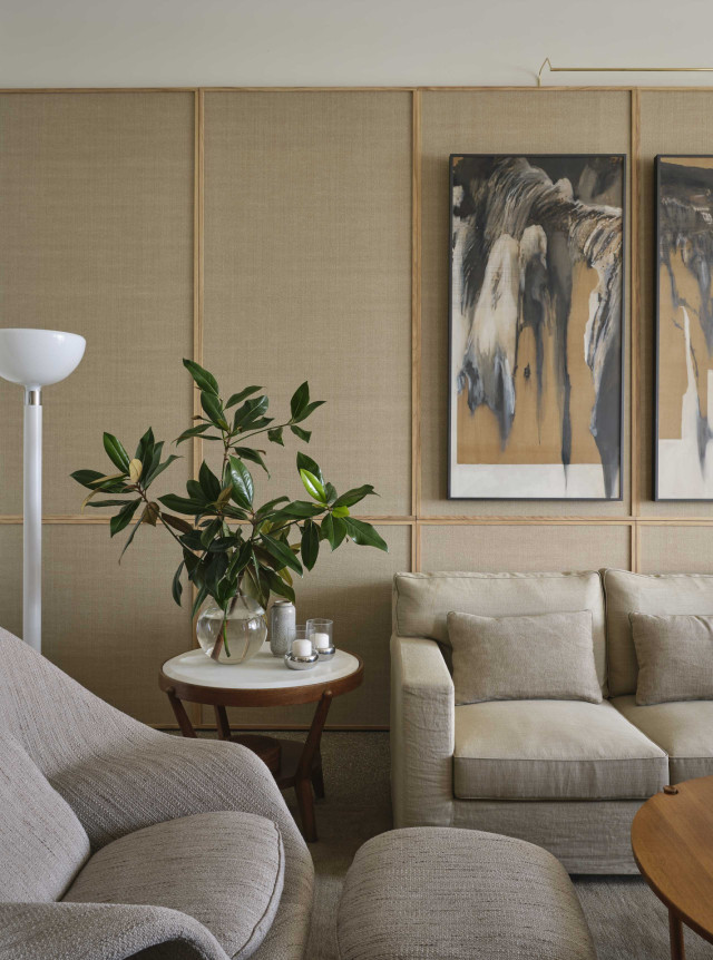 A cozy living room features a beige sofa and armchair. A round wooden side table holds a plant and candles. Two abstract paintings hang on a paneled wall behind.