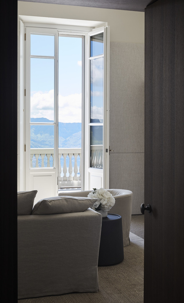 A comfortable seating area with grey chairs faces an open balcony door. Beyond, a scenic view of mountains and a blue sky with clouds is visible, providing a serene atmosphere.