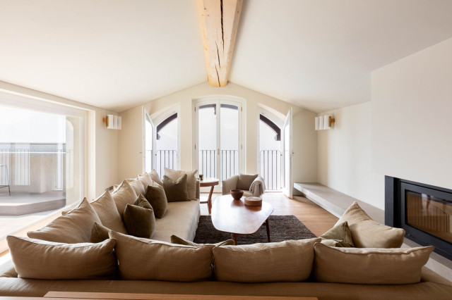 A spacious living room features a large, beige sectional sofa facing a wooden coffee table with decorative items. The room is well-lit by natural light from two open French doors leading to a balcony.