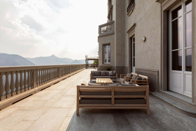 Outdoor furniture set on a spacious stone terrace, overlooking mountain views and bordered by a balustrade, adjacent to a large, elegant building.