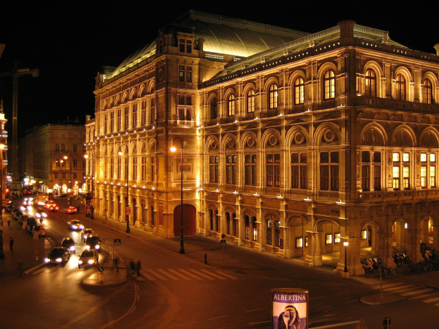 An illuminated, ornate building stands lit at night, with cars driving on a city street nearby