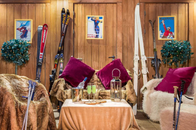 Ski equipment rests against wooden walls adorned with vintage posters. Fur-draped chairs and a table with drinks create a cozy, alpine setting. Two small decorated trees flank the scene.