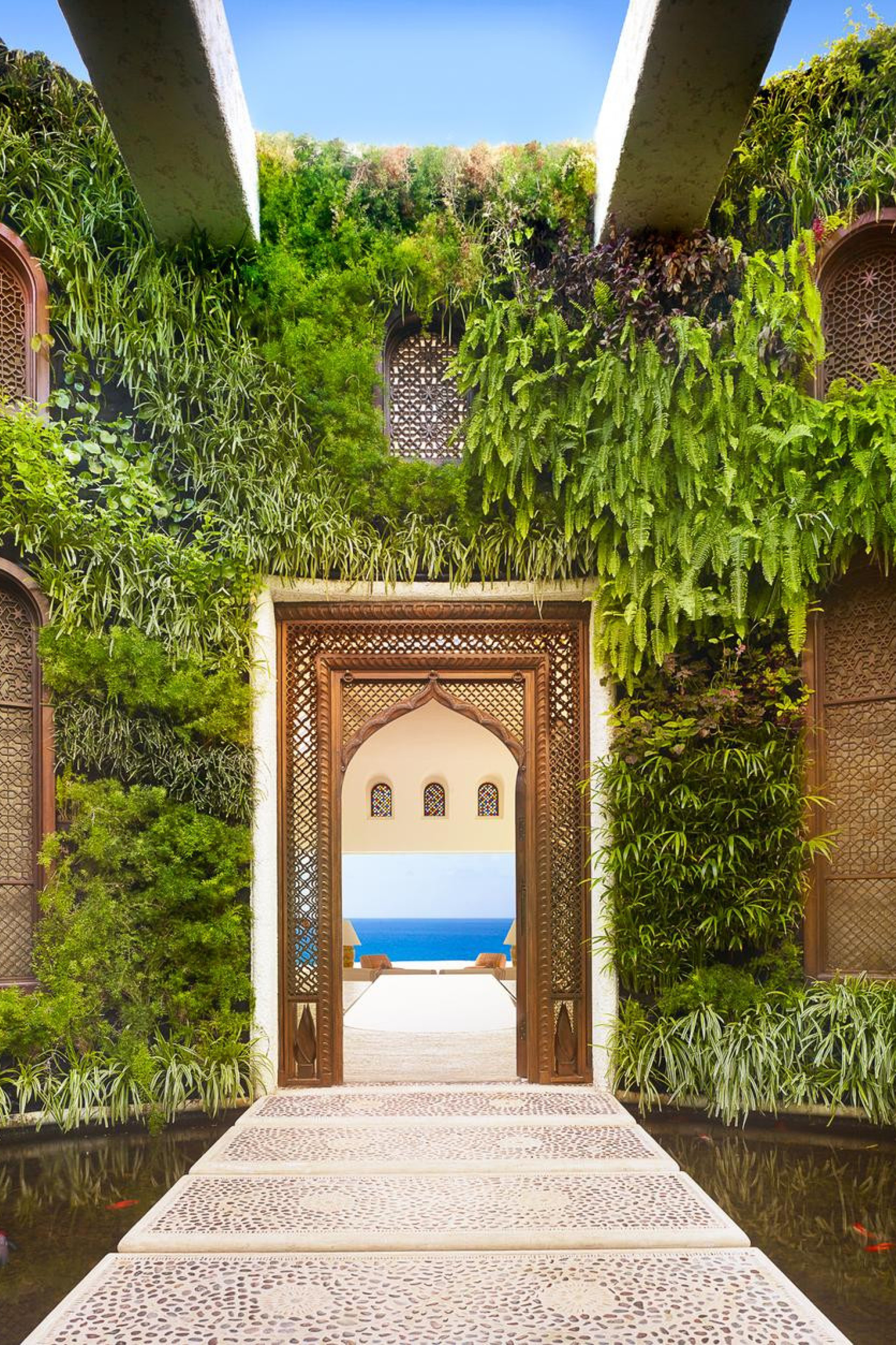 An ornate archway leads to an open balcony overlooking blue seas. Surrounding lush vertical gardens with intricate windows frame the scene, under a bright blue sky.